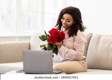 Holidays And Gift Delivery. Happy Black Young Woman Holding Bouquet Of Red Roses And Showing To Webcam, Sitting On Couch At Home And Using Laptop, Having Video Call. Virtual Celebration Concept - Powered by Shutterstock