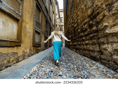 Holidays in Italy. Blonde woman wearing beige sweater, turquoise skirt walking with hands spread out along cobblestone narrow street in old town of Bergamo in autumn. Back view. Sightseeing old city. - Powered by Shutterstock