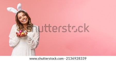Holidays, spring and party concept. Portrait tender, romantic blond girl in white dress and rabbit ears, tilt head cute, smiling happy as holding painted eggs, celebrating Easter, pink background