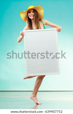 Holidays summer and advertisement concept. Woman wearing yellow hat and bikini holding blank presentation board. Female model posing in full length on blue background.