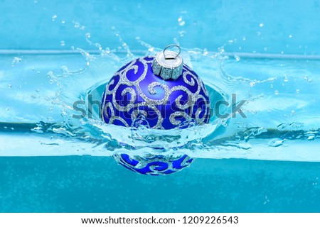 Holidays and vacation concept. Festive decoration for Christmas tree, blue ball dropped into water with splashes, blue background. Christmas decoration or toy for Christmas tree swim in pool.