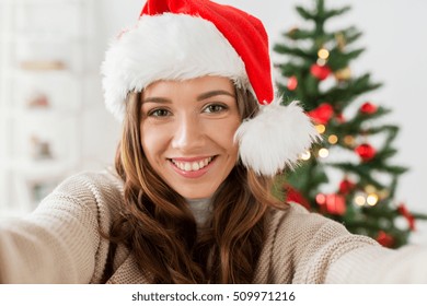 holidays, winter and people concept - happy young woman in santa hat taking selfie over christmas tree at home