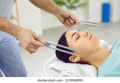 Holistic Doctor Working With Female Patient. Specialist Holding Two Resonating Tuning Forks Near Young Woman's Head. Close Up Of Healer's Hands. Sound Therapy, Stress Reduction, Relaxation Concept