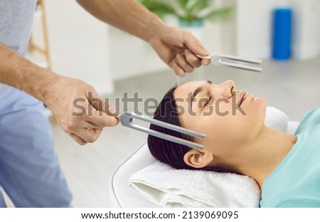 Holistic doctor working with female patient. Specialist holding two resonating tuning forks near young woman's head. Close up of healer's hands. Sound therapy, stress reduction, relaxation concept