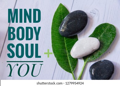 Holistic Health Concept. Top View Of Zen Stones With Green Leaves On White Wooden Table. Text Mind Body Soul You. 