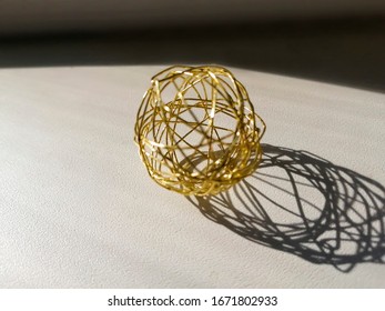 a hollow sphere woven from gilded wire lies with shadow on a light textured surface.