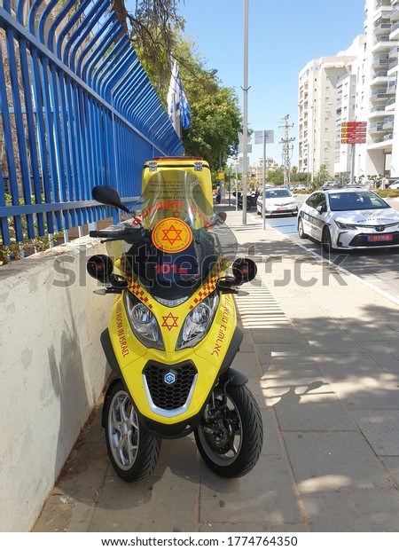 HOLON,\
ISRAEL. July 12, 2020. Yellow Israel Magen David Adom First Aid\
paramedic scooter bike and a police car parked on the side of the\
road. Israel emergency services concept\
image.