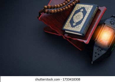 Holy Al Quran with written arabic calligraphy meaning of Al Quran, lantern lamp and rosary beads or tasbih on black background.