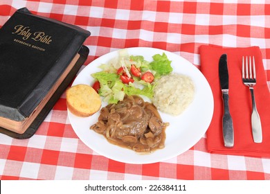 Holy Bible On Red Plaid Tablecloth  -- Sunday Potluck Dinner Of Salisbury Steak In Brown Gravy With Onions And Mushrooms On White Plate With Mashed Potatoes And Tossed Salad And Cornbread Muffin.