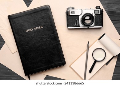 Holy Bible with photo camera, magnifier, notebook and paper sheets on dark wooden background