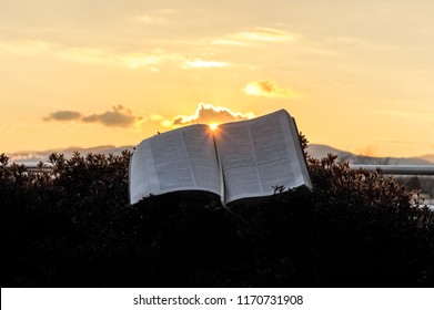 The Holy Bible (Word Of God) Opened During A Glorious Golden Sunset. Sun In The Center Of The Bible. Background With Clouds And Bright Sun And Intense Orange And Gold Color. Horizontal Shot.