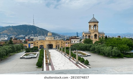 Holy Trinity Cathedral complex opens across a formal courtyard in Tbilisi, with its ceremonial gate, bell tower, fountains, and Mtatsminda skyline supporting pilgrimage and cultural heritage tourism.