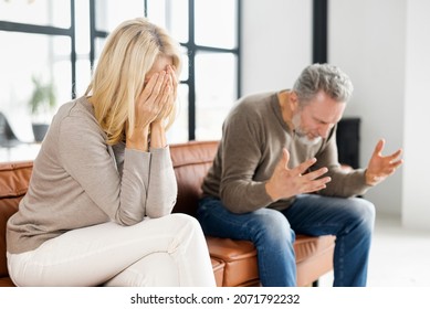 Home Abuse Concept. Depressed Mid-aged Woman Sits On The Couch And Crying, An Irritated And Angry Mature Man Is Screaming On The Background. A Couple Is Arguing At Home