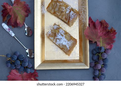 Home baked apple strudel. Sliced apple roll on golden plate with spoons on black background. Apple strudel slices sprinkled with sugar powder. Traditional autumn and winter dessert.