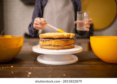 Home baker assembling layered sponge cake with frosting brush and glass of water in warm kitchen