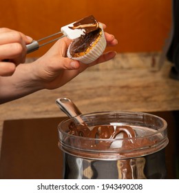 Home Baking: Hands Of A Woman Covering With A Kitchen Scraper A Cupcake Fresh From The Oven With Melted Chocolate With A Pot And A Bowl And A Woody Background.