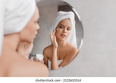 Home beauty routine. Young woman applying moisturising cream on face looking at miror in bathroom, free space. Attractive lady wearing white towel on head after shower - Powered by Shutterstock