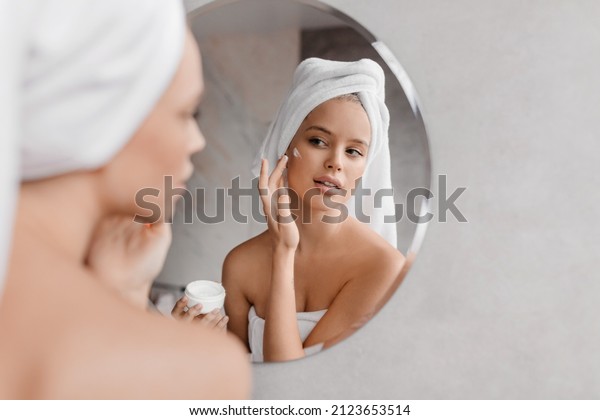 Home beauty routine.\
Young woman applying moisturising cream on face looking at miror in\
bathroom, free space. Attractive lady wearing white towel on head\
after shower