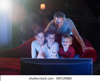 At Home By Night, Cheerful Family Sitting In A Red Couch And Watching A Funny Movie On Tv. They Are Laughing Togetherness. Shot With Flare