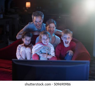At home by night, Cheerful family sitting in a red couch and watching a funny movie on tv. They are laughing togetherness. Shot with flare