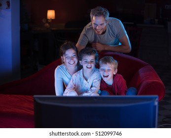 At Home By Night, Cheerful Family Sitting In A Red Couch And Watching A Funny Movie On Tv. They Are Laughing Togetherness