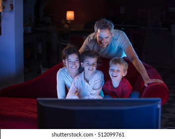 At Home By Night, Cheerful Family Sitting In A Red Couch And Watching A Funny Movie On Tv. They Are Laughing Togetherness