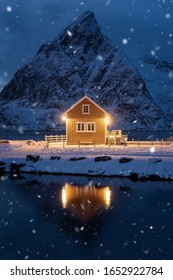 Home, Cabin Or House At Night, Norwegian Fishing Village In Reine City, Lofoten Islands, Nordland County, Norway, Europe. White Snowy Mountain Hills, Nature Landscape Background In Winter Season.