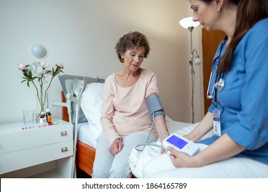 Home Caregiver Checking Blood Pressure Of Senior Patient Sitting On Hospital Bed. Home Care Nurse Taking Blood Pressure Of Senior Woman With Digital Blood Pressure Monitor In Bedroom.