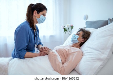 Home Caregiver Comforting Senior Female Patient Lying In Bed. Side View Of Nurse Wearing Surgical Protection Mask Talking To Elderly Female Patient At Hospice.