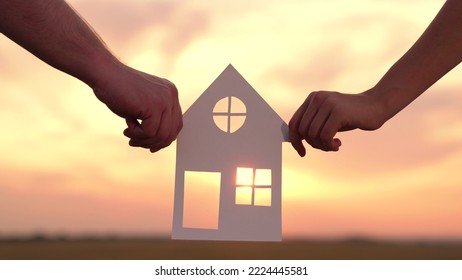 Home For Children And Parents, Familys Hands Are Holding Paper House At Sunset, Sun Is Shining Through Window. Symbol Of House, Happiness. Concept Of Building House For Family. Dream To Buy House.