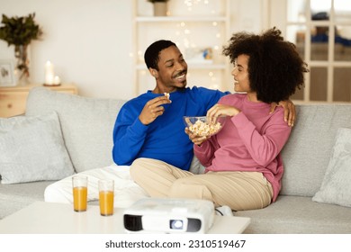 At Home Cinema. Happy Black Millennial Couple Watching Film Enjoying Movie Night Together, Eating Popcorn Sitting On Sofa Near Projector In Living Room Indoor. Modern Entertainment. Selective Focus - Shutterstock ID 2310519467