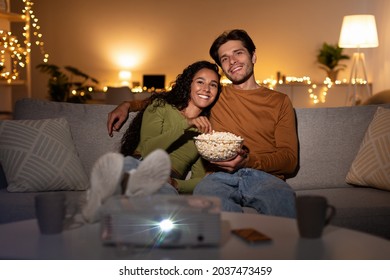 Home Cinema. Happy Couple Watching Movie Using Projector Eating Popcorn And Relaxing Enjoying Evening Sitting On Couch In Living Room Indoors. Cheerful Spouses Watch Film Together. Family Lifestyle