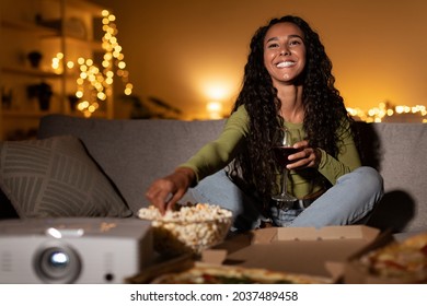 Home Cinema. Happy Woman Watching Movie Using Projector, Drinking Wine And Eating Popcorn Sitting On Couch In Living Room Indoors. Entertainment And Fun, Weekend Leisure Concept