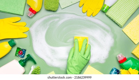 Home cleaning products. A hand in a green rubber glove wipes soap suds with a washcloth on a green background. Concept of cleanliness, cleaning services. - Shutterstock ID 2186239999