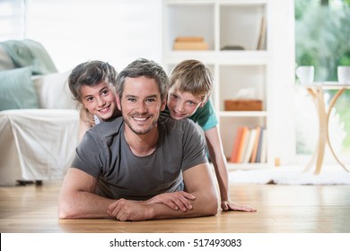 At Home, Dad And His Two Kids Lying On Wooden Floor In The Living-room. They Are Smiling While Looking At Camera