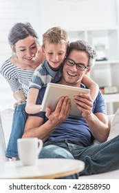 At home, Dad, mom and their young son having fun by gaming together on a tablet, they are sitting on a white couch in the living room and the boy looks at the screen over the shoulder of his father
