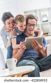 At home, Dad, mom and their young son having fun by gaming together on a tablet, they are sitting on a white couch in the living room and the boy looks at the screen over the shoulder of his father - Shutterstock ID 422858953