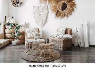 Home decor concept. Comfortable wicker furniture, rattan armchair with cushions, bamboo coffee table and macrame on white wall in cozy living room with ethnic interior design