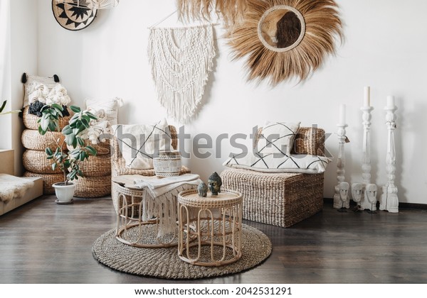 Home decor\
concept. Comfortable wicker furniture, rattan armchair with\
cushions, bamboo coffee table and macrame on white wall in cozy\
living room with ethnic interior\
design