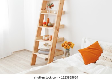 Home Decor. Cozy Fall Bedroom Interior: White Wall, Bed With White Linen, Light Beige Plaid, Orange Pillows, Wooden Rack, Vase With Yellow Chrysanthemums Flowers, Pumpkins, Candles. Autumn Decoration.