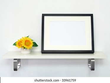 Home Decoration Picture Frame. Potted Sunflowers And A Black Picture Frame On White Shelf Against A White Wall.