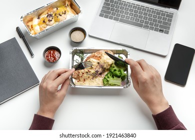 Home Delivery During Covid-19 Lockdown And Health Meal. Hands Of Man With Knife And Fork, Guy Eats Food From Container On White Table, Near Notepad, Laptop And Smartphone With Empty Screen, Pov