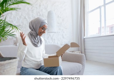 Home delivery of mail. Young Muslim woman in hijab received parcel, happy sitting on sofa and opening box, online shopping, received gift. - Powered by Shutterstock