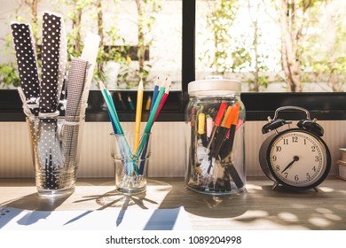 Home Design Idea - Creative Thinking Study Room Working Table Decorating Items With Clock, Colorful Pens And Pencils In Jar And Glass In Front Of Sunlight Black Aluminium Window At Warming Daytime