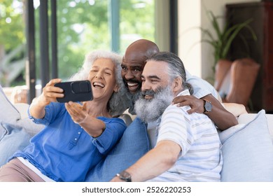 At home, diverse senior friends taking selfie, laughing together. Sitting closely on couch with a blue background, enjoying each other company, sharing joyful moments, unaltered - Powered by Shutterstock