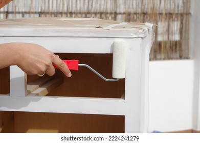 home DIY: a woman's hand with a gold ring holds a small roller with white paint while restoring a pine wood chest of drawers, protected with masking tape - Shutterstock ID 2224241279