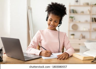 Home Education Concept. Black Girl Using Laptop Computer Doing Homework Online Wearing Headset Sitting At Desk Indoor - Shutterstock ID 1803080557