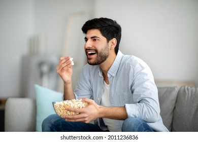 Home Entertainments. Excited Arab Guy Eating Popcorn And Watching TV, Spending Free Time At Home. Happy Man Enjoying Movie While Relaxing On Couch In Living Room, Free Space