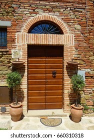 Home Entrance With A Brown Wooden Door With Black Knocker, Set In A Stone Doorway With An Arched Window Overhead, With Matching Shrubs In Terra Cotta Pots And Empty Flower Pots And A Small Welcome Mat