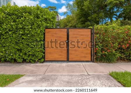 Home entrance driveway gate with wood slats in Miami, Florida. Gate of a residence in the middle of bush walls with concrete sidewalk and driveway.